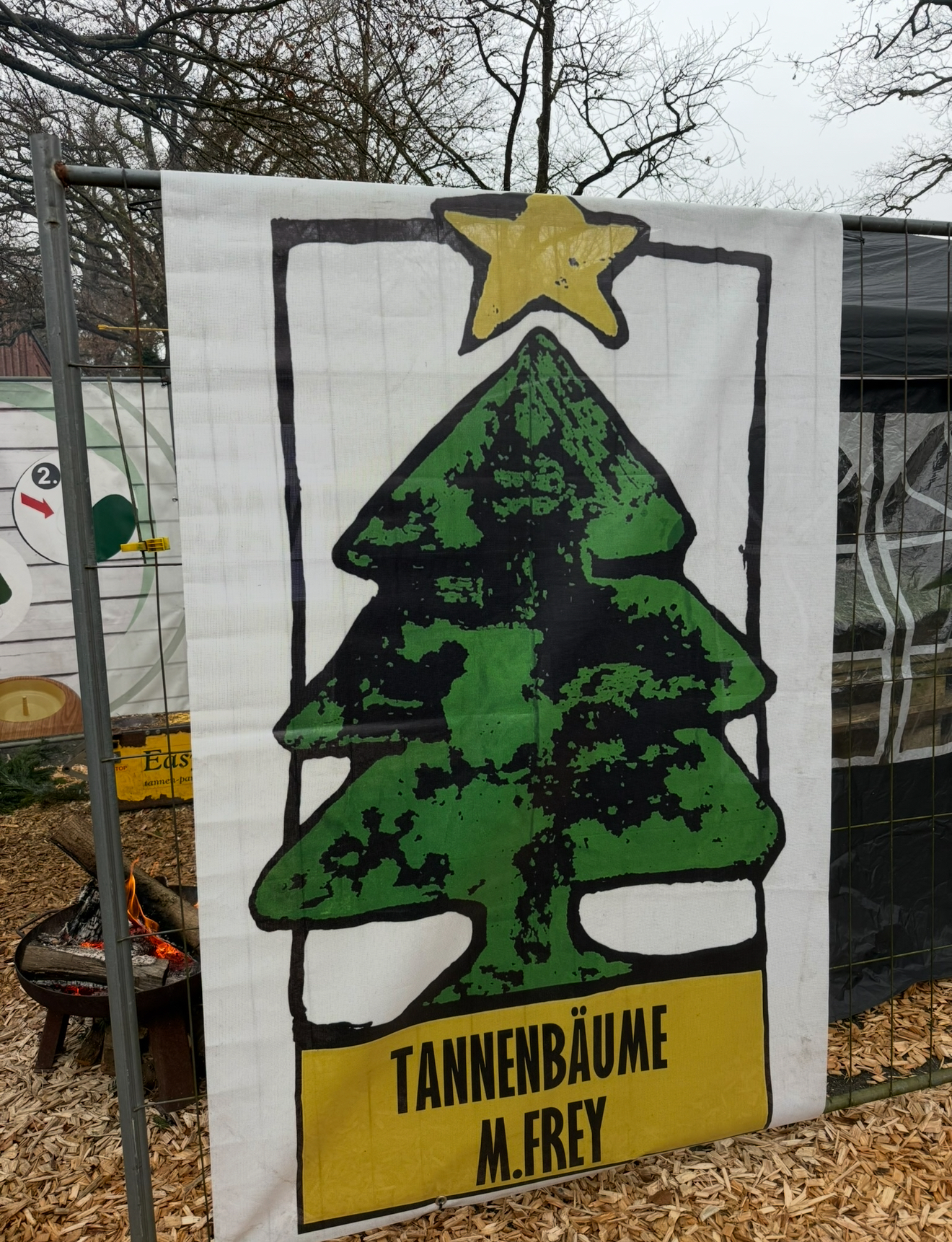 Weißes Banner mit grüner Tanne, gelbem Stern und der Aufschrift TANNENBÄUME M. FREY, befestigt am Bauzaun des Verkaufsstands neben einer brennenden Feuerschale