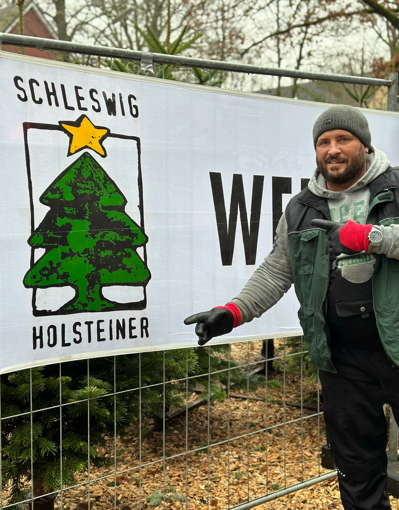 Manuel Frey in grauer Strickmütze und grüner Arbeitsweste zeigt lächelnd mit beiden Fingern auf das weiße Schleswig-Holsteiner-Banner mit grüner Tanne und gelbem Stern